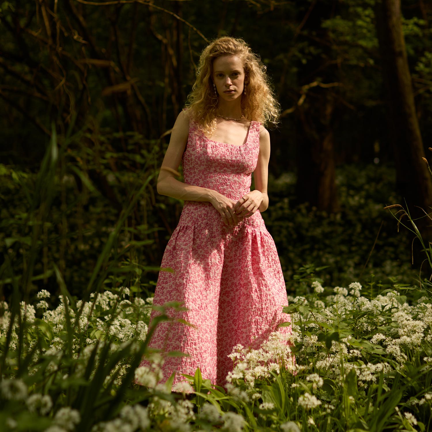 A person in a pink dress stands in a lush forest surrounded by white flowers.