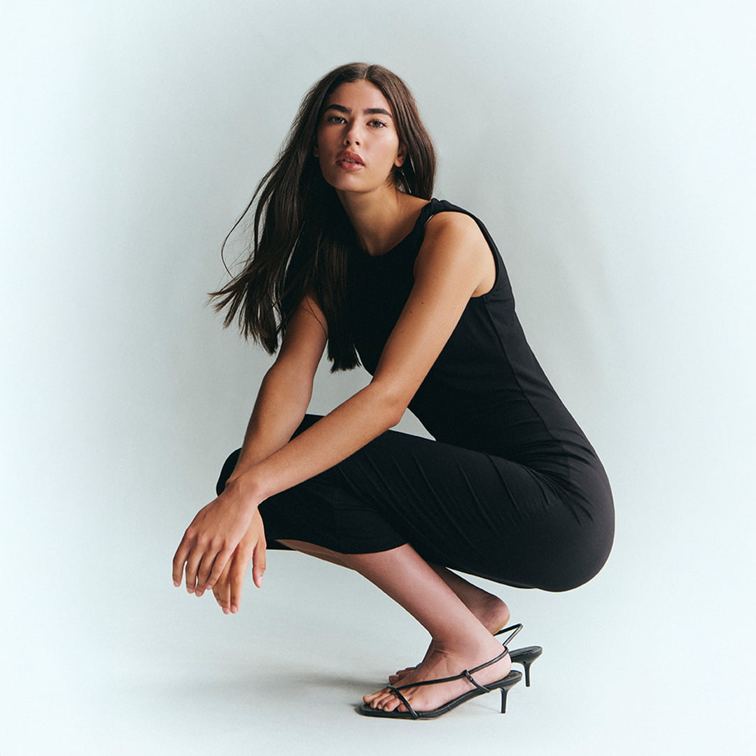 Woman in a black dress and heels crouching on a plain background.