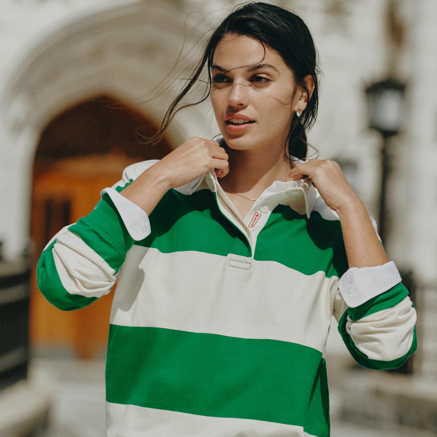 Woman in a green and white striped shirt and jeans, standing outside with blurred architectural background.