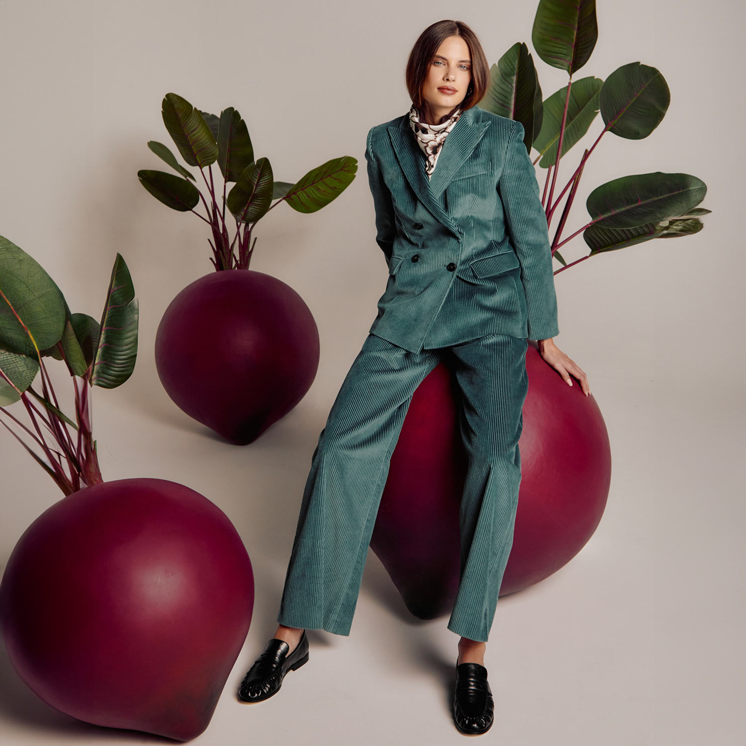 A person in a teal suit leans against oversized abstract beet-like sculptures with green leaves.