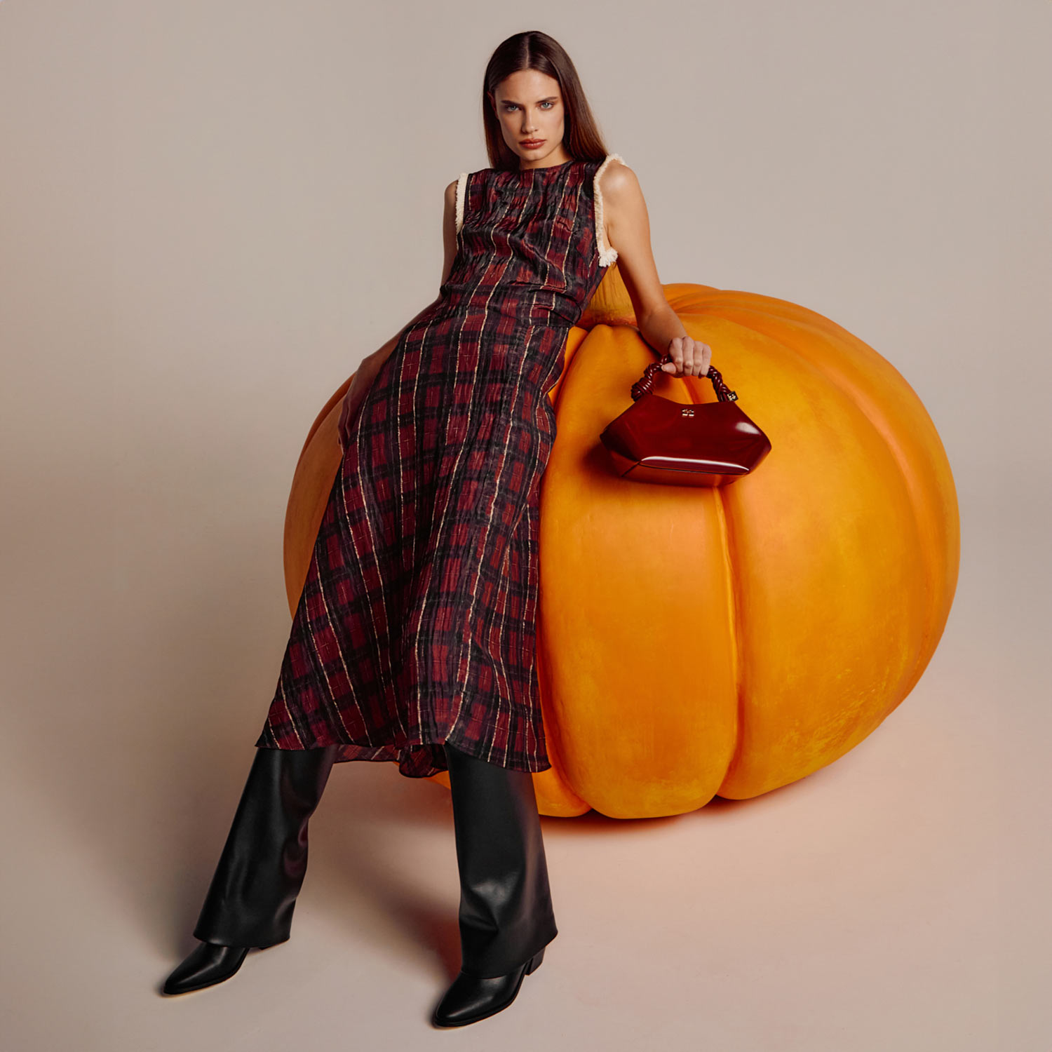 A woman in a plaid dress and black boots leans against a large pumpkin, holding a red handbag.