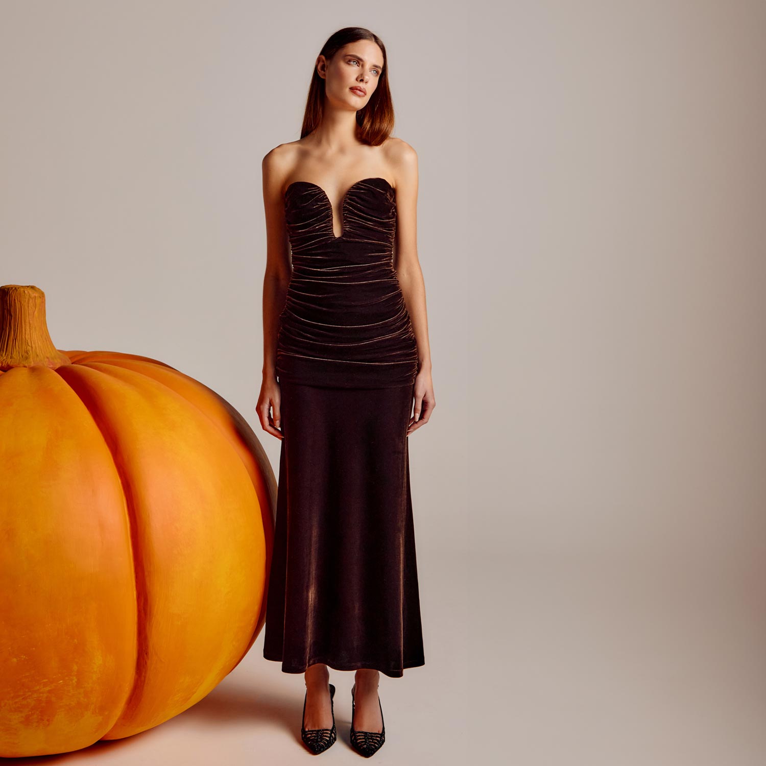 A woman in a strapless black dress stands next to a giant pumpkin.