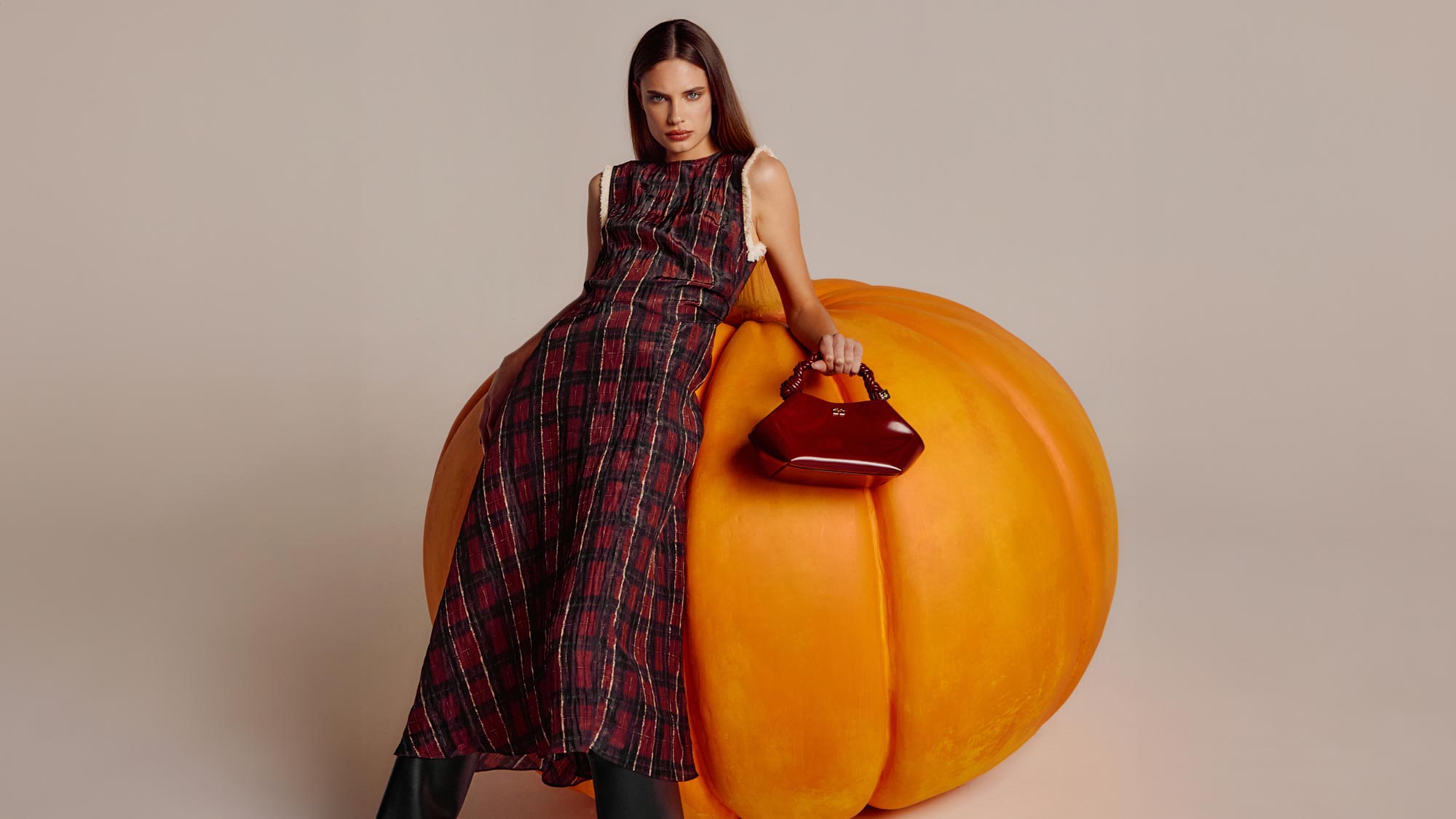 A woman in a plaid dress leans on a large pumpkin while holding a brown handbag.