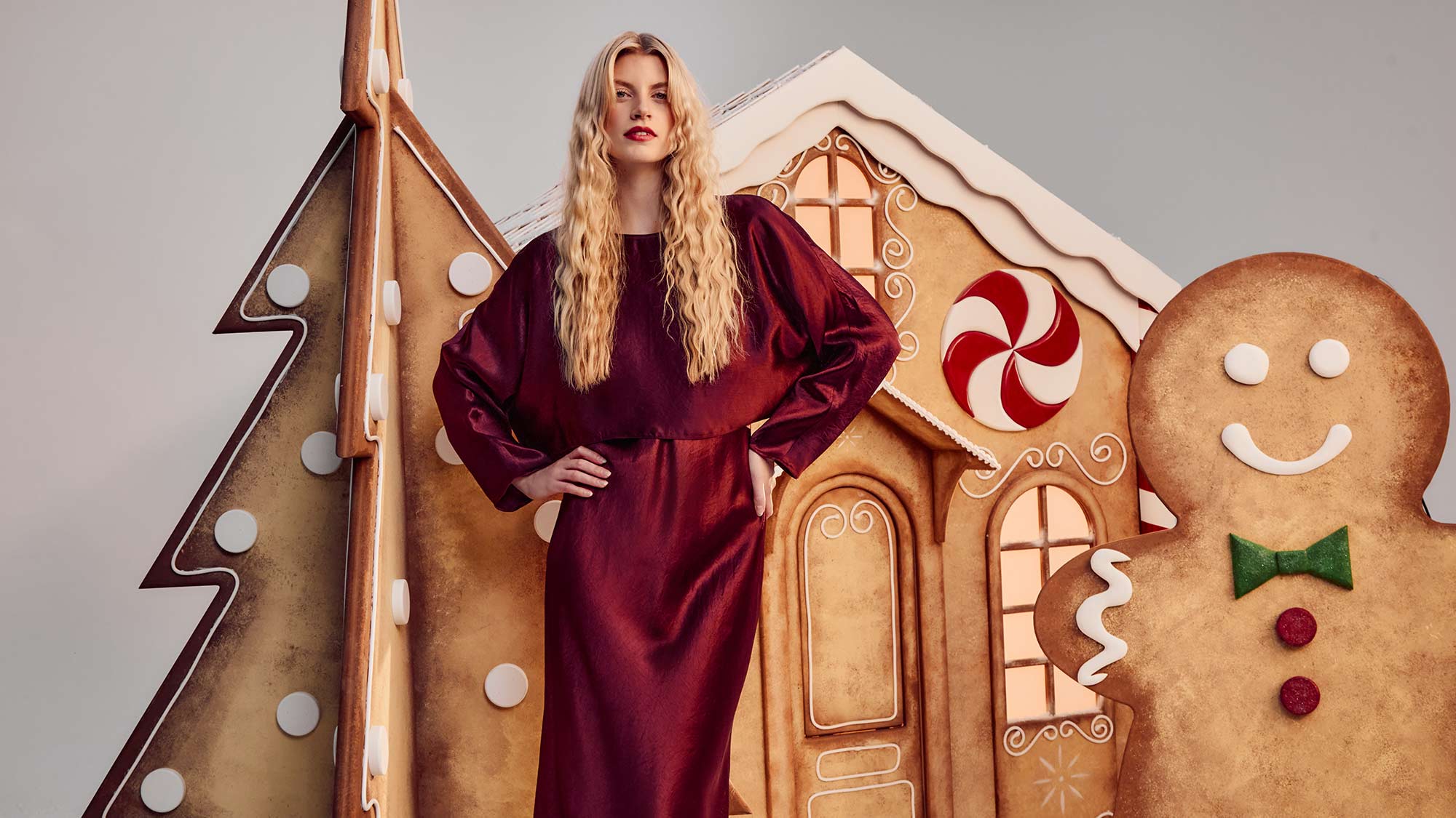 Woman in a burgundy dress stands by large gingerbread decorations, including a house and gingerbread man.