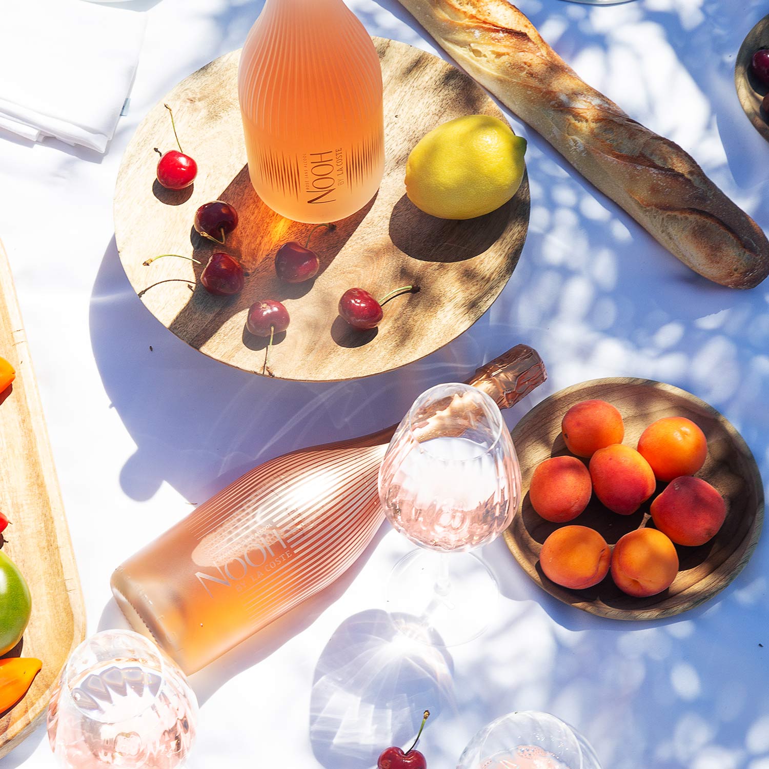A bottle of ros&eacute; wine on a table with glasses, cherries, and apricots, set under tree dappled sunlight.