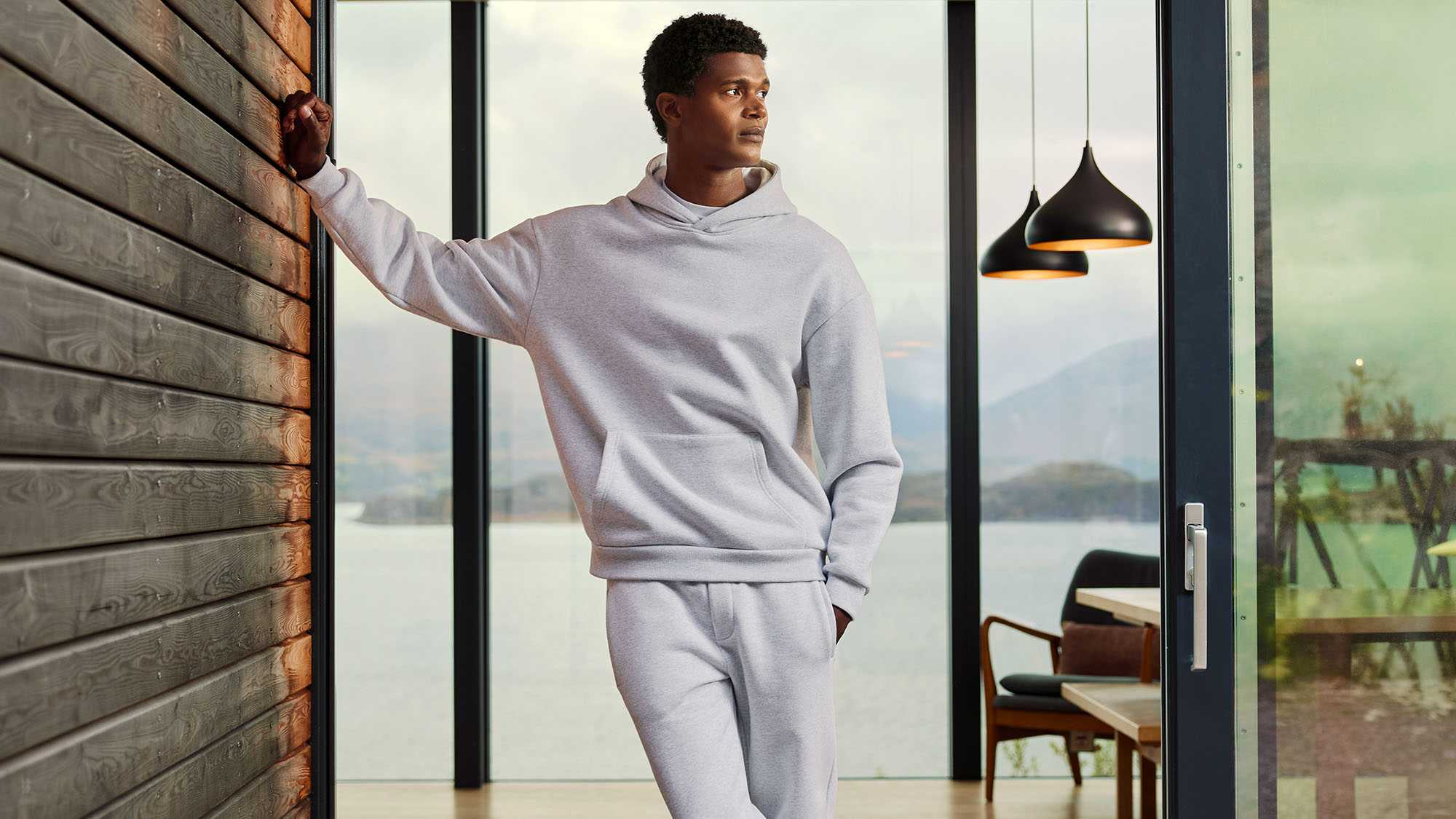 Man in grey marl hoodie and matching joggers standing by wooden wall, overlooking scenic lake and mountain views
