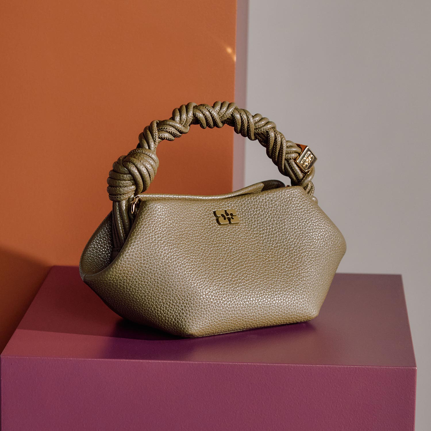 Gold textured leather handbag with knotted rope handles on coloured block display against neutral background