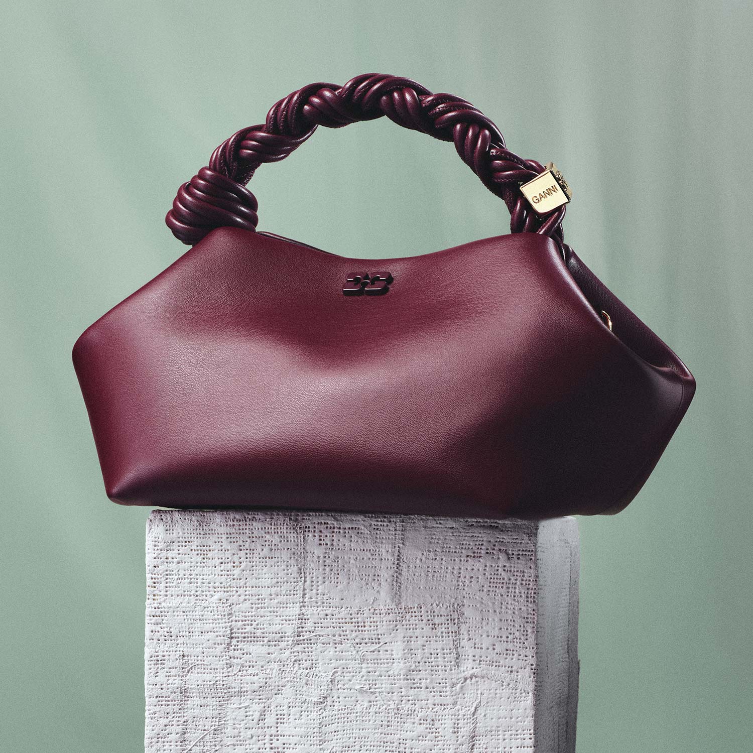 A burgundy handbag with a twisted handle sits on a textured stone block against a soft green background.
