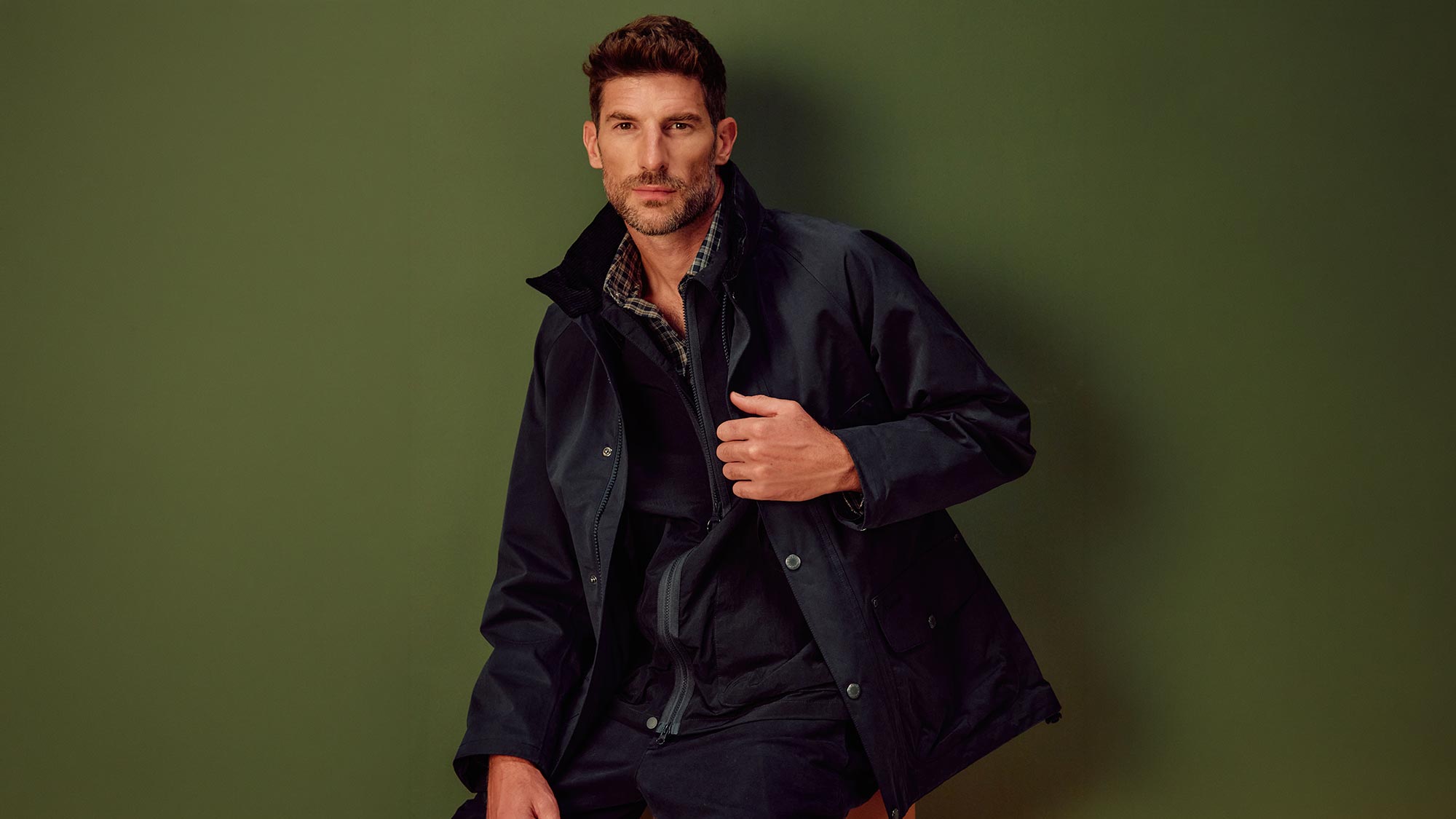 Man wearing navy waxed jacket with tartan shirt against sage green background
