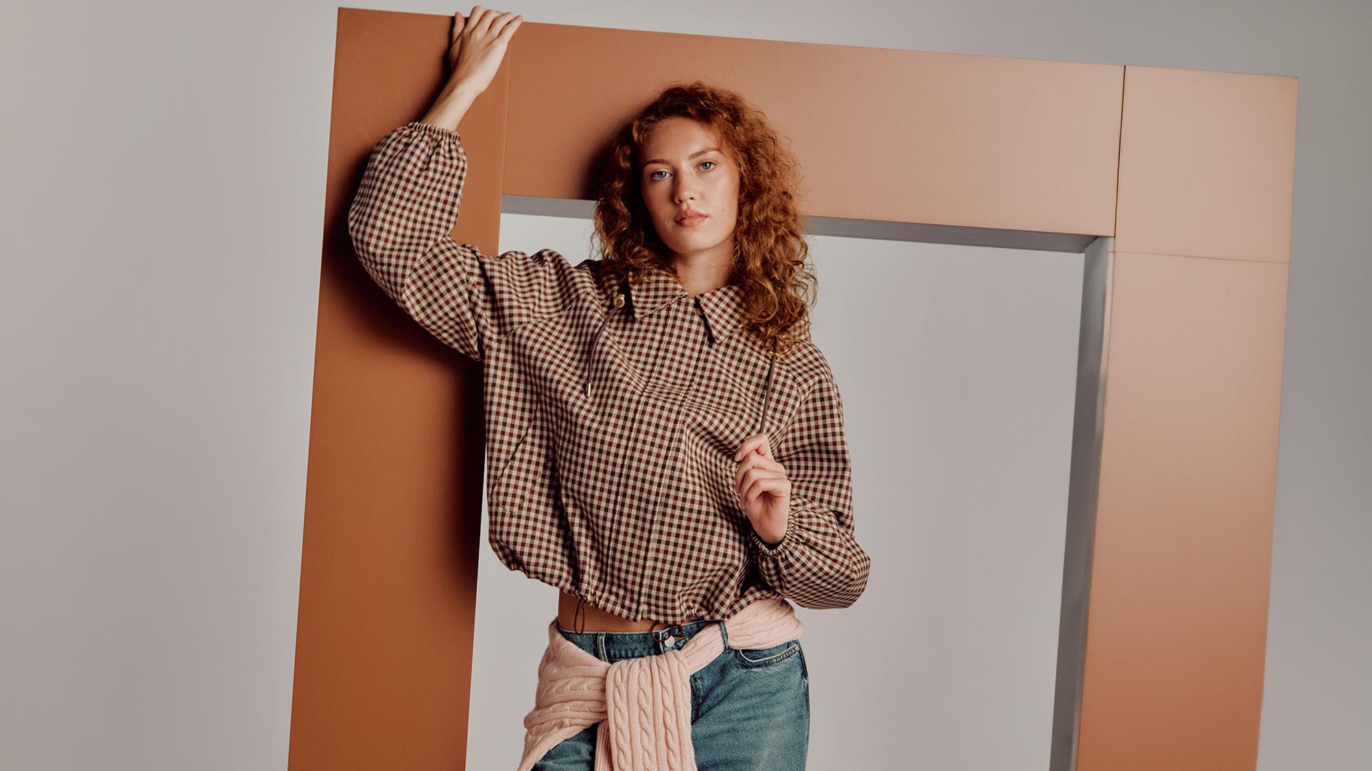 Woman wearing brown gingham shirt and cream cable knit cardigan against geometric brown and white backdrop