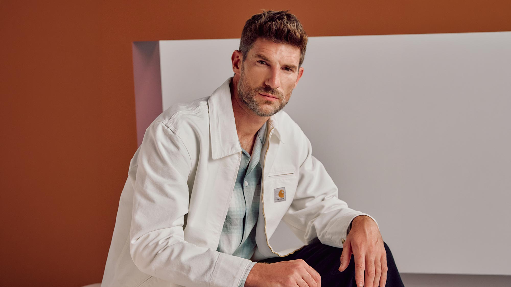 Man wearing cream Carhartt jacket over blue shirt against brown and grey background