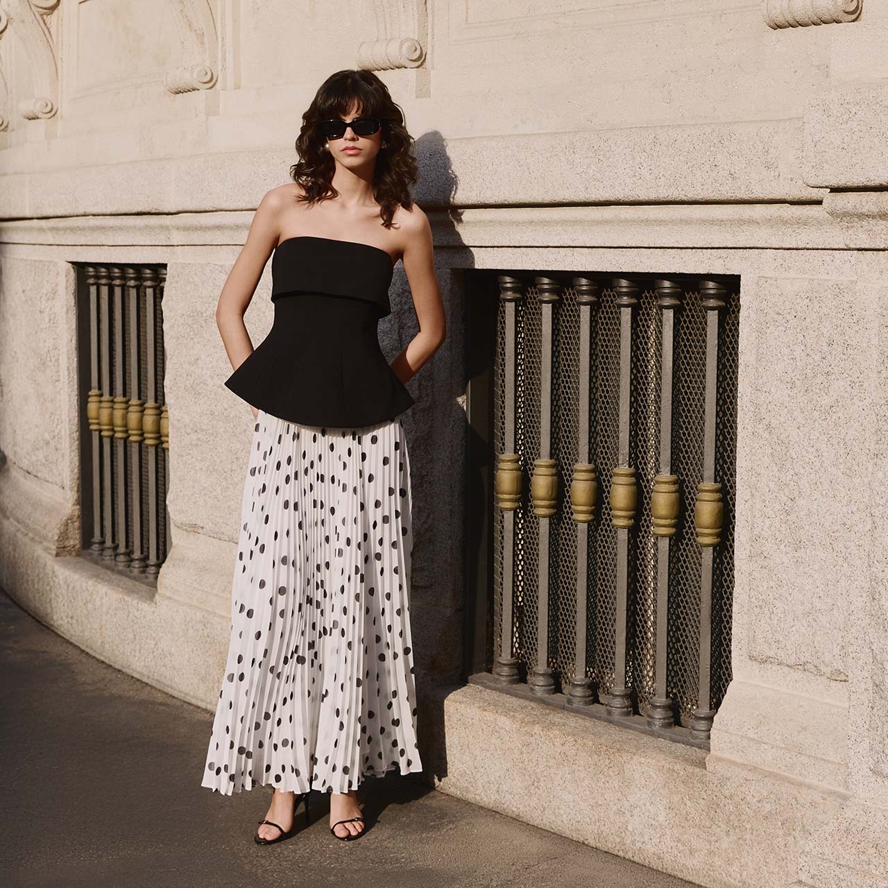 Woman wearing black strapless top and white pleated polka dot skirt with dark sunglasses