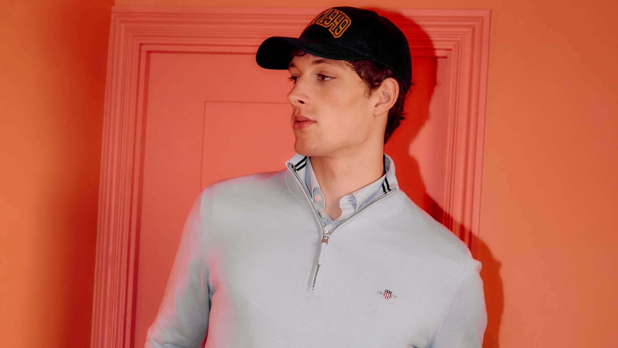 A person wearing a light sweater and cap stands in front of a reddish-orange wall.