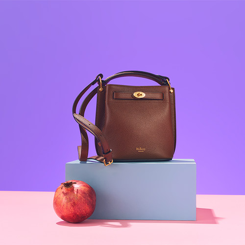 A brown leather handbag on a blue platform with a pomegranate on a pink and purple background.