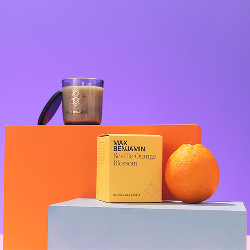 Candle, yellow box labeled 'Max Benjamin Seville Orange Blossom,' and an orange on colorful blocks against a purple background.