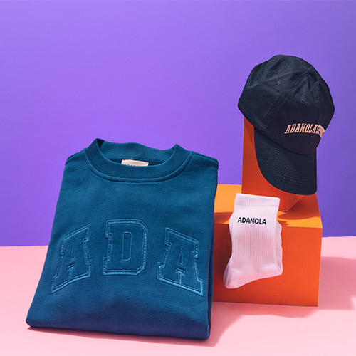 A folded teal sweatshirt, white socks, and a black cap with 'ADANOLA' displayed against a purple background.