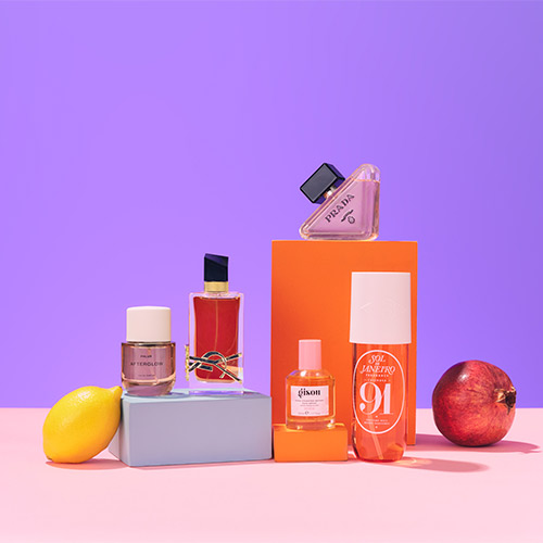 Perfume bottles arranged with a lemon and a pomegranate on a pink and purple background.