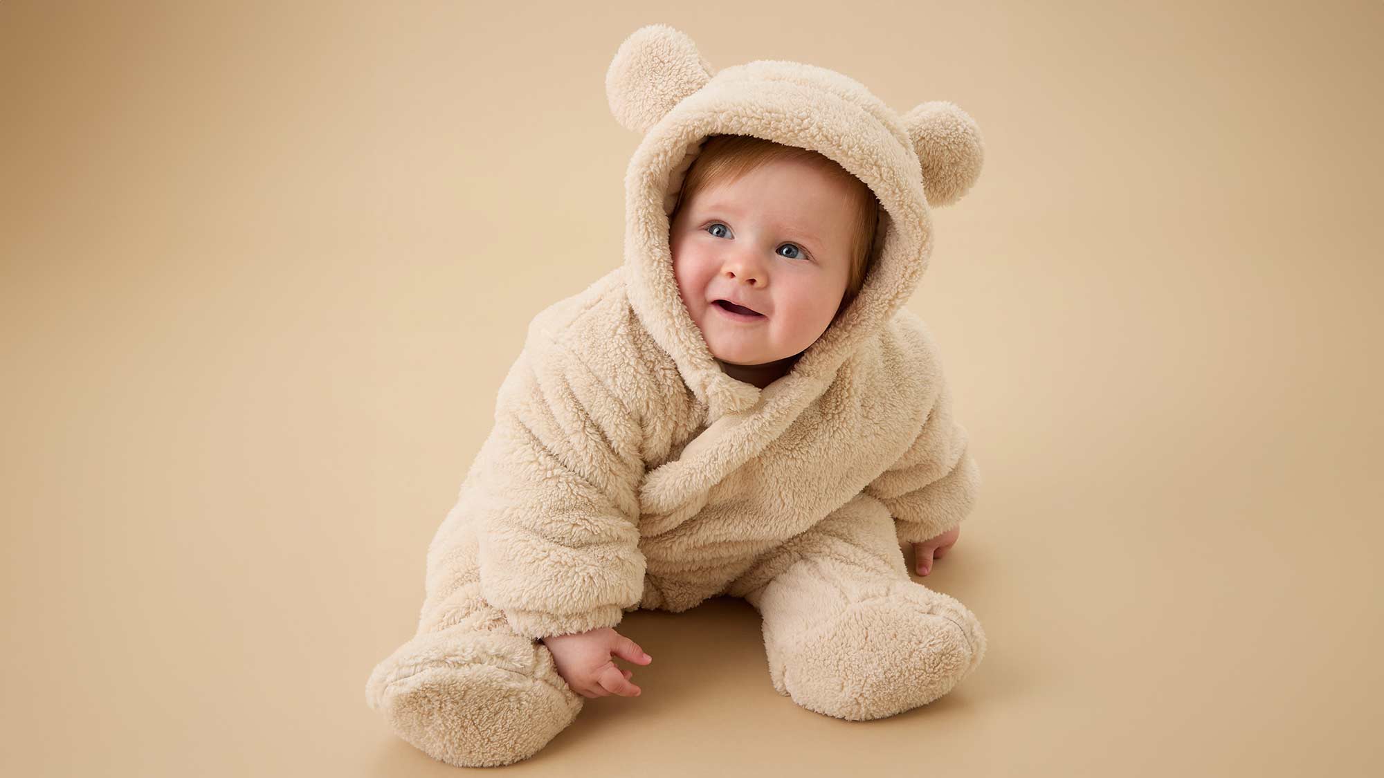 A baby in a fluffy, beige bear costume with ears sits on the floor smiling.