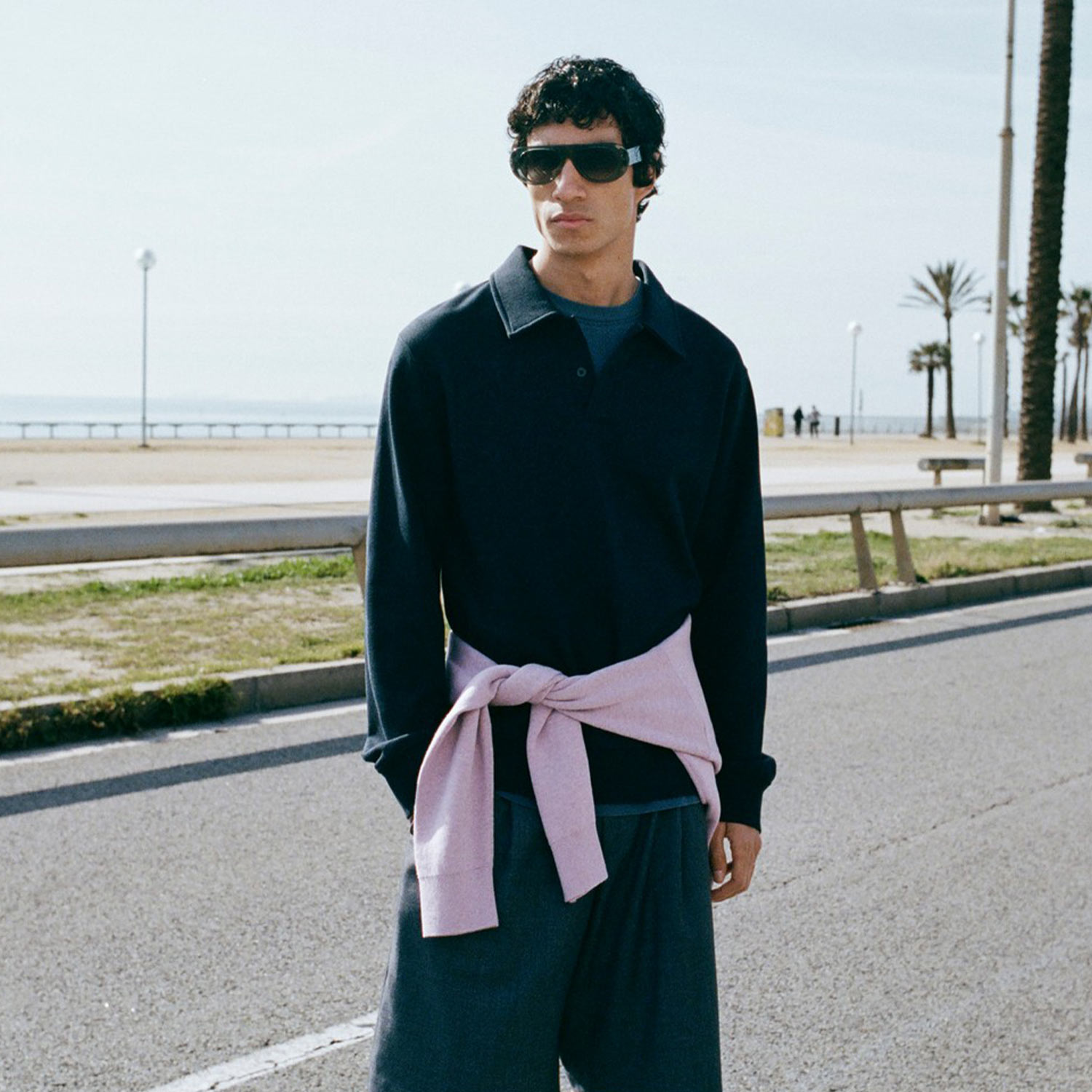 Man at beach wearing black polo shirt and trousers with pink tied jumper around waist and dark sunglasses