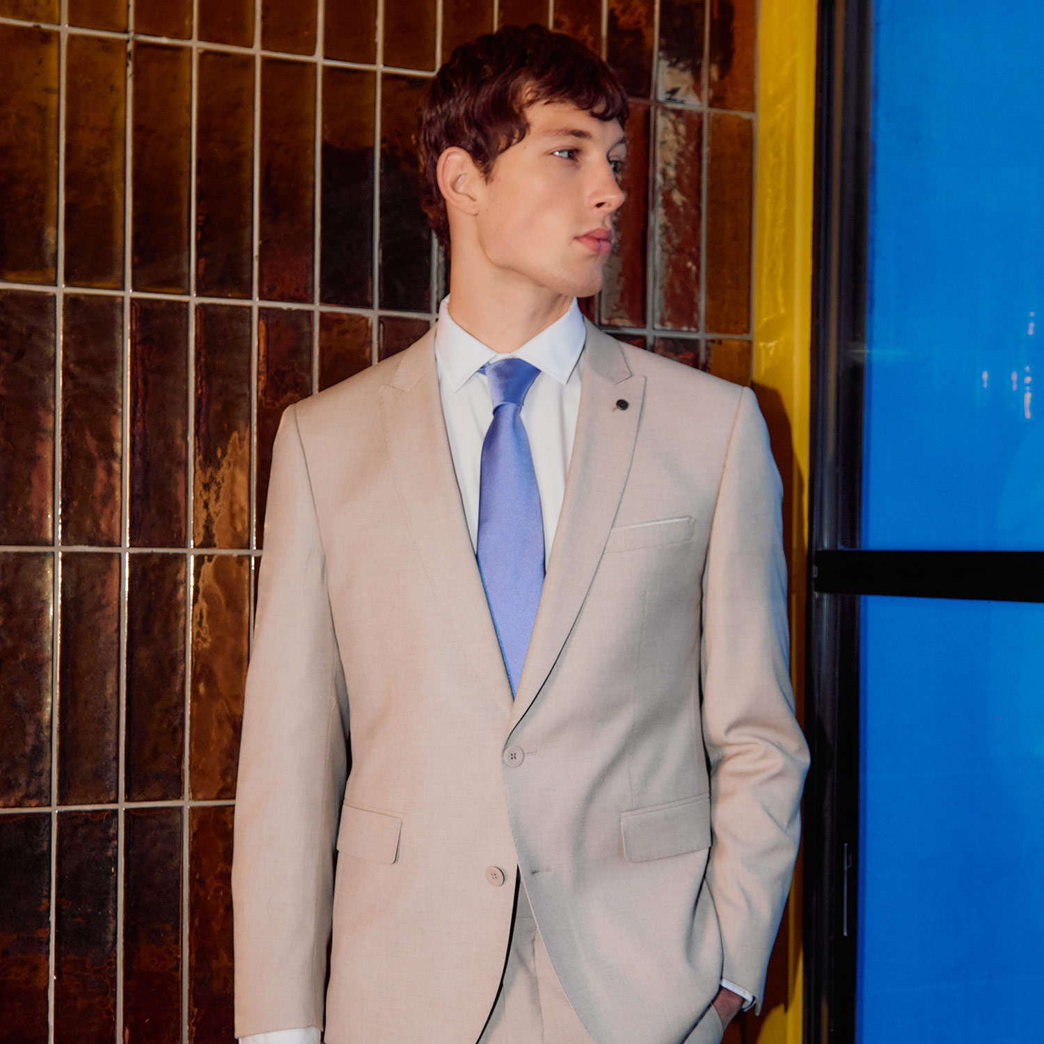 A person in a beige suit with a blue tie stands in front of a textured wall.