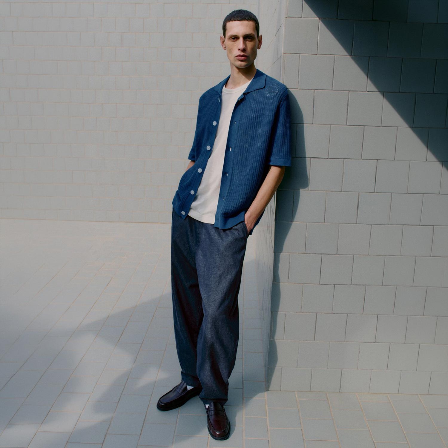 Man wearing blue short-sleeved shirt with white t-shirt and dark denim trousers, standing against white tiled wall