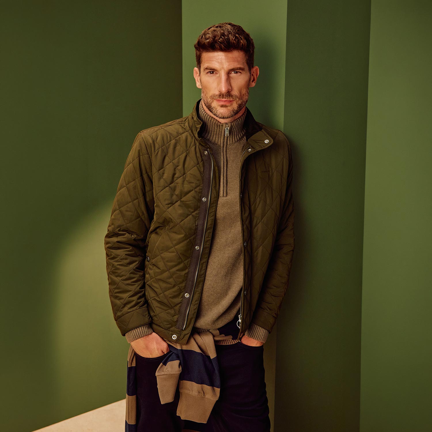 Man wearing olive quilted jacket over beige roll-neck jumper and patterned trousers against green background