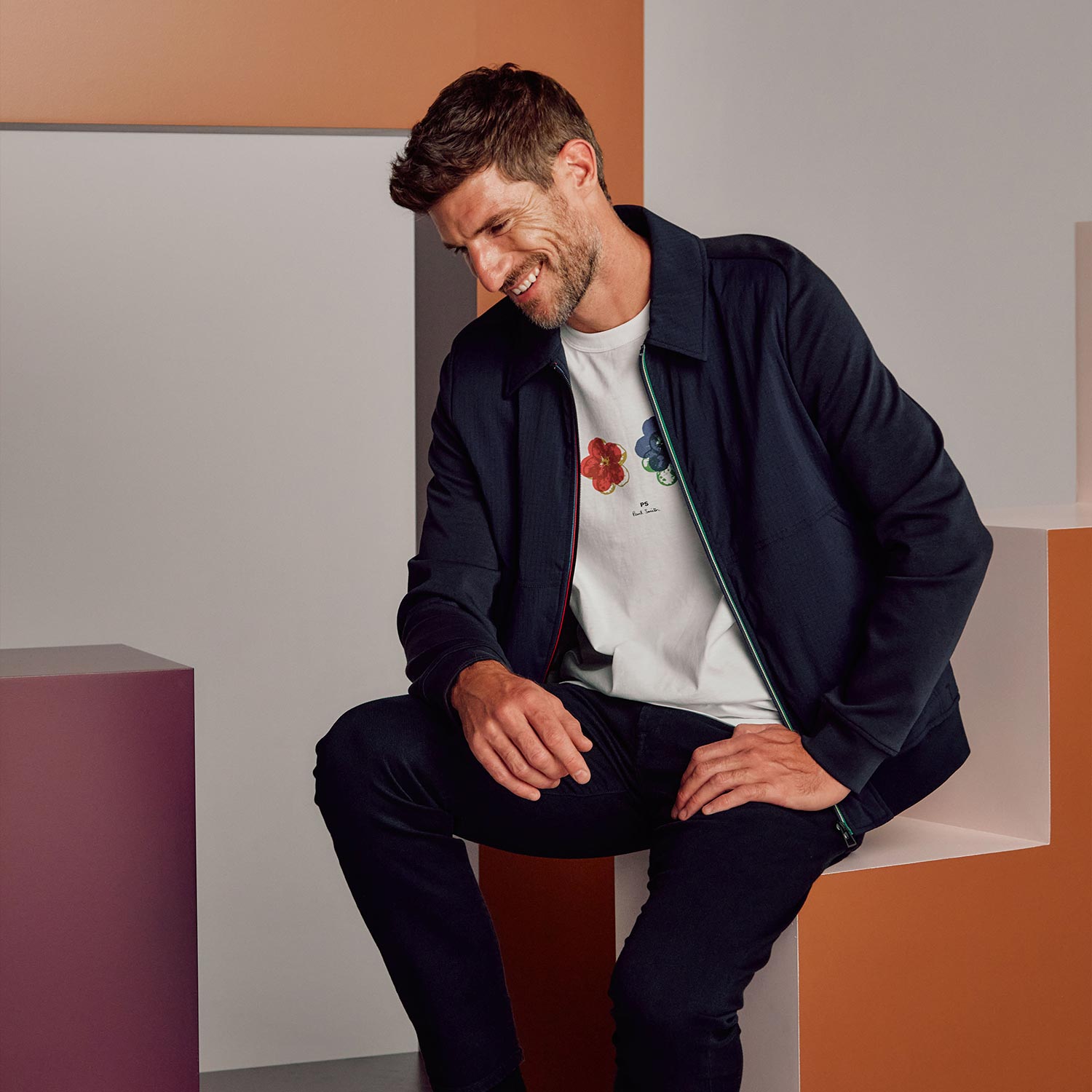Man wearing navy jacket and graphic t-shirt seated against geometric coloured background
