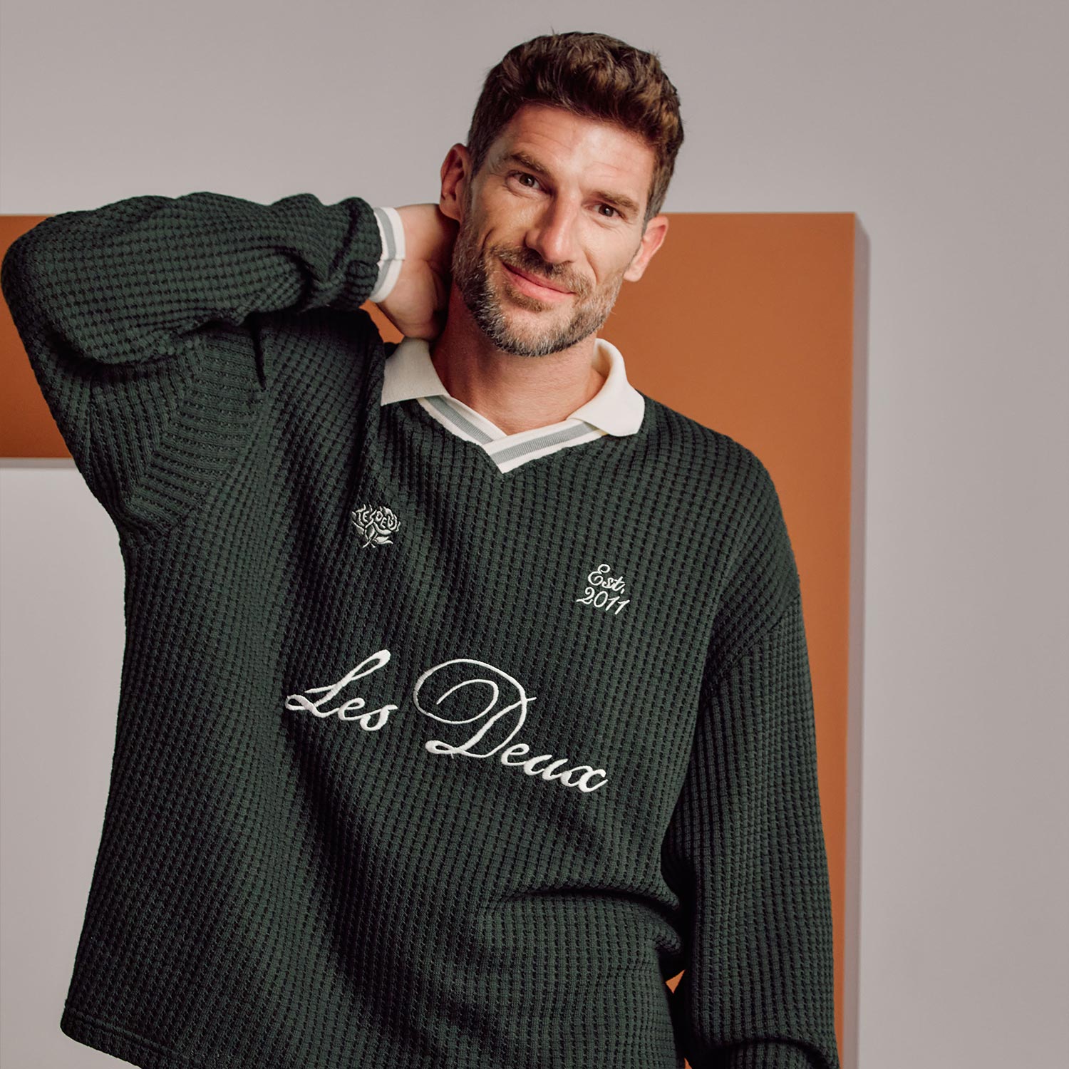 Man wearing forest green ribbed jumper with embroidered text and white collar detail against orange background