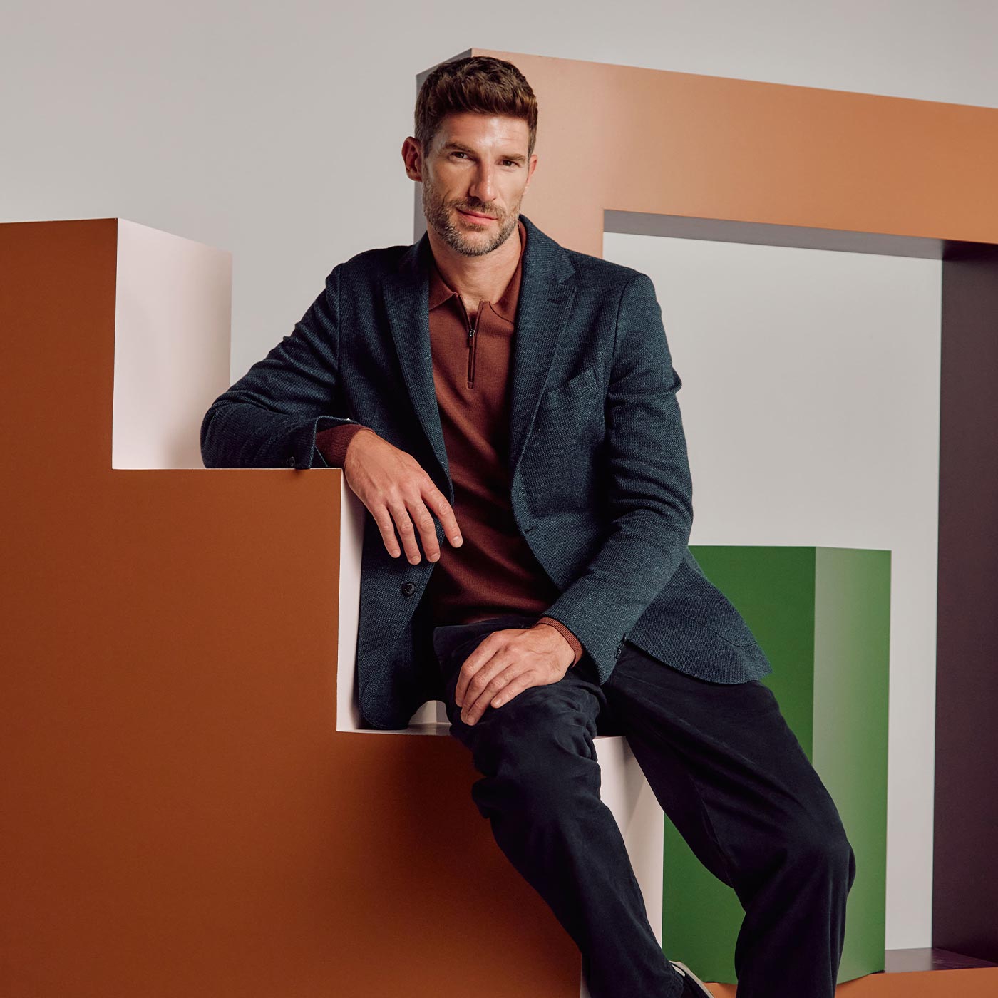 Man wearing charcoal blazer, burgundy shirt, and navy trousers seated against colourful geometric background