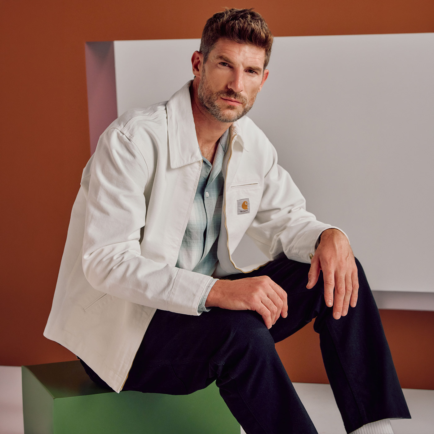 Man wearing cream overshirt with blue check shirt and navy trousers, seated against geometric background