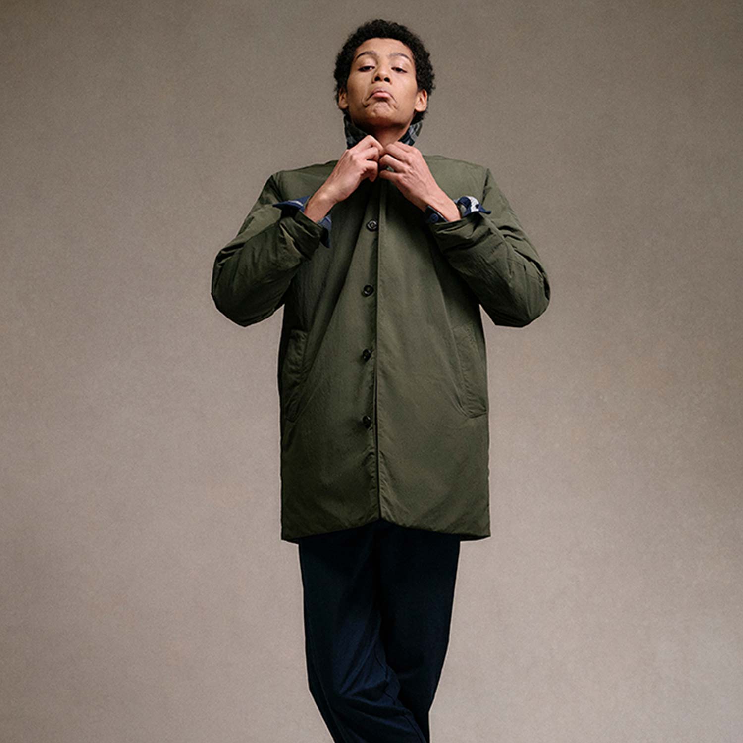 Person wearing a green coat and dark pants, standing with legs crossed and hands near the collar, in front of a neutral background.