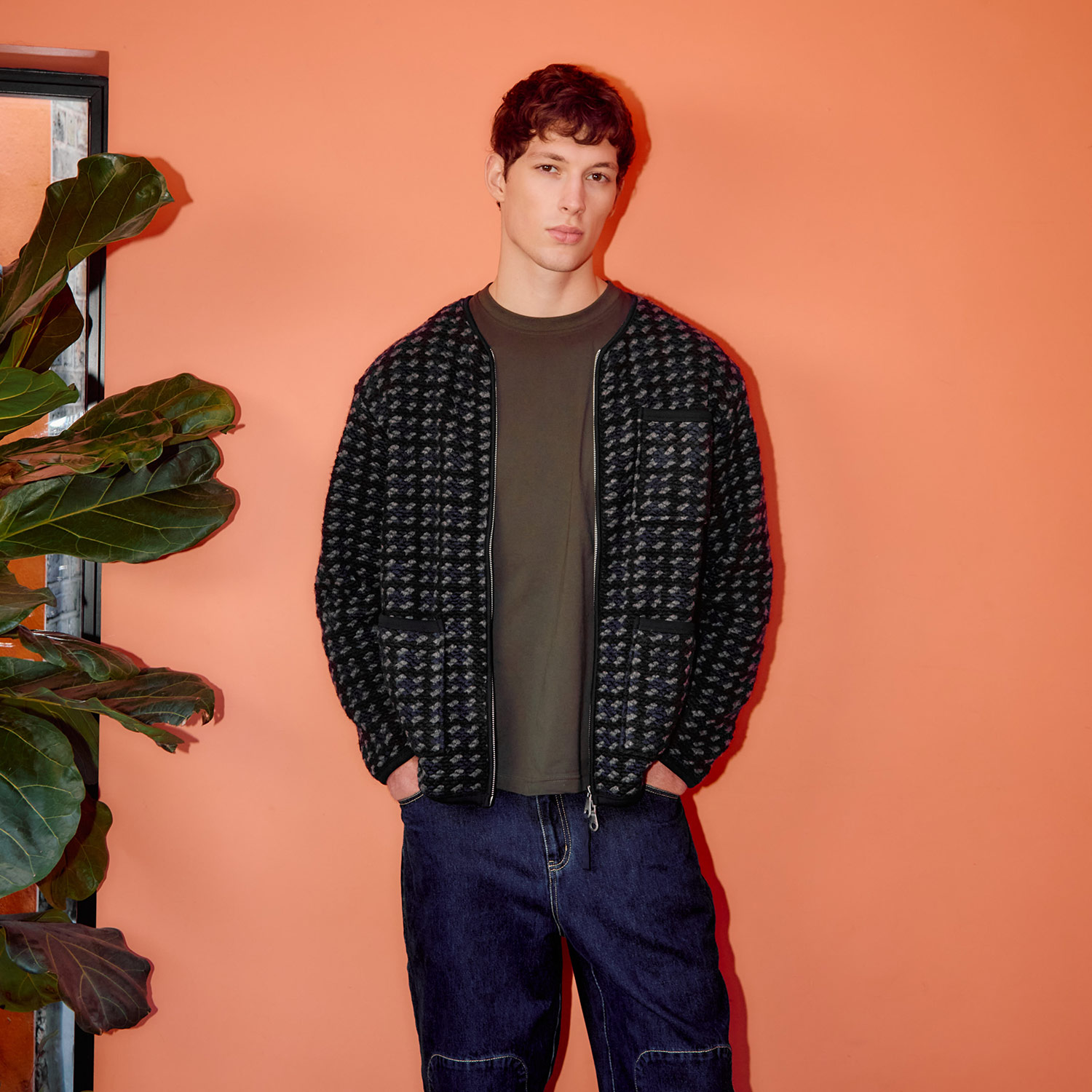 A person stands against a peach-colored wall, wearing a patterned jacket and dark shirt, next to a large green plant.