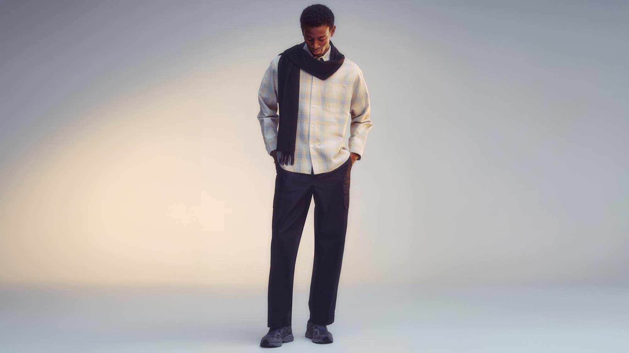 A person in a plaid shirt and dark pants stands with a scarf draped around their neck against a soft-lit background.