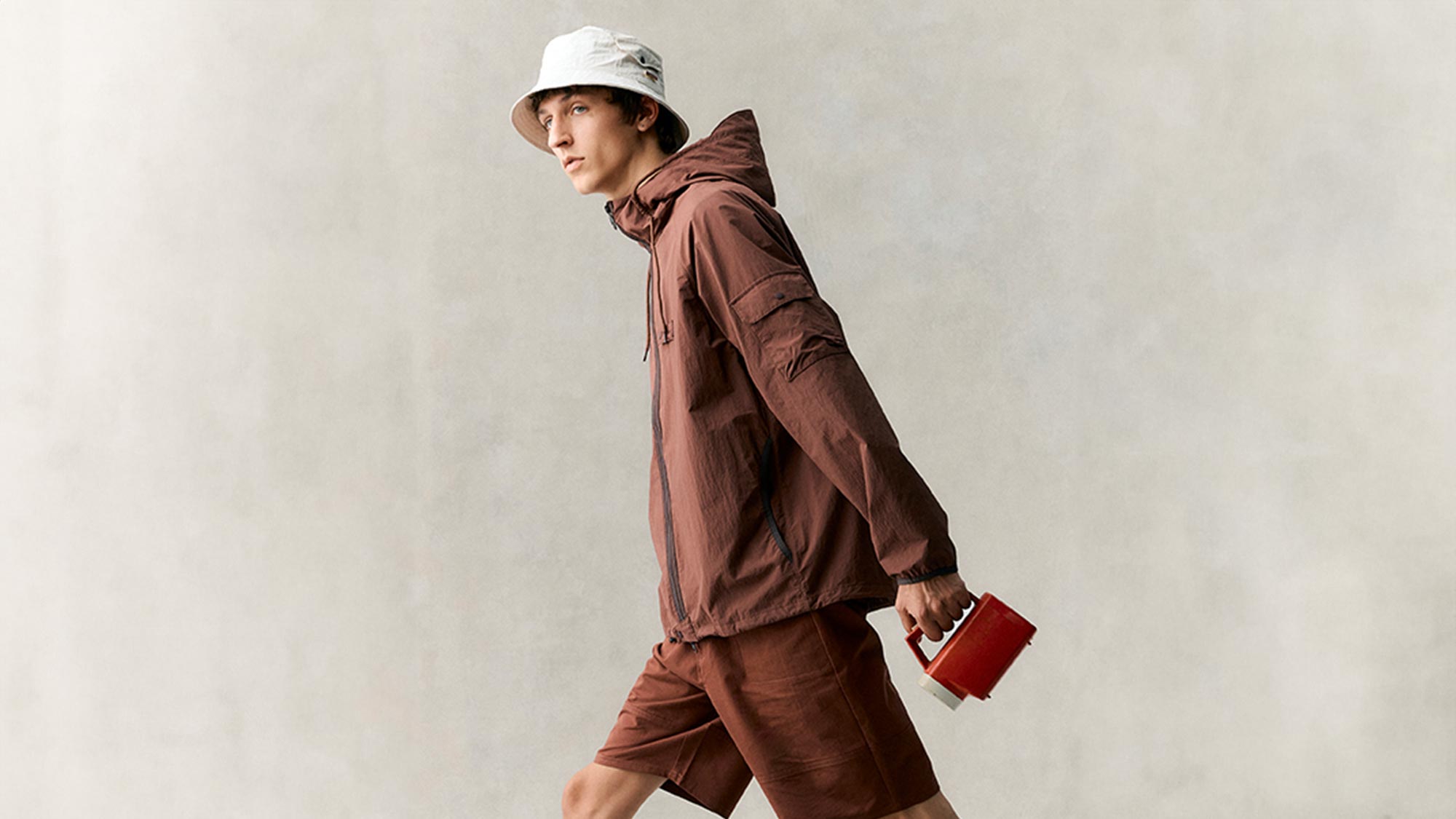 Man wearing burgundy hooded jacket and shorts with white bucket hat, holding red leather wallet