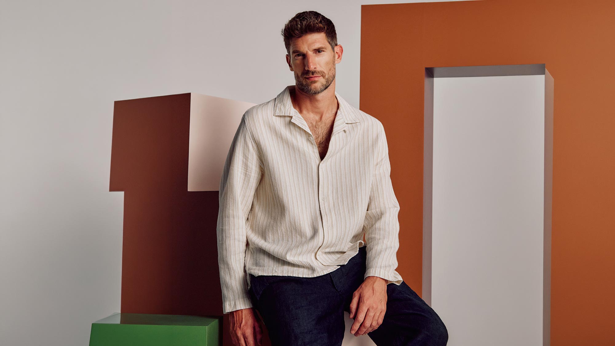 Man wearing cream striped shirt and navy trousers seated against geometric orange and white background