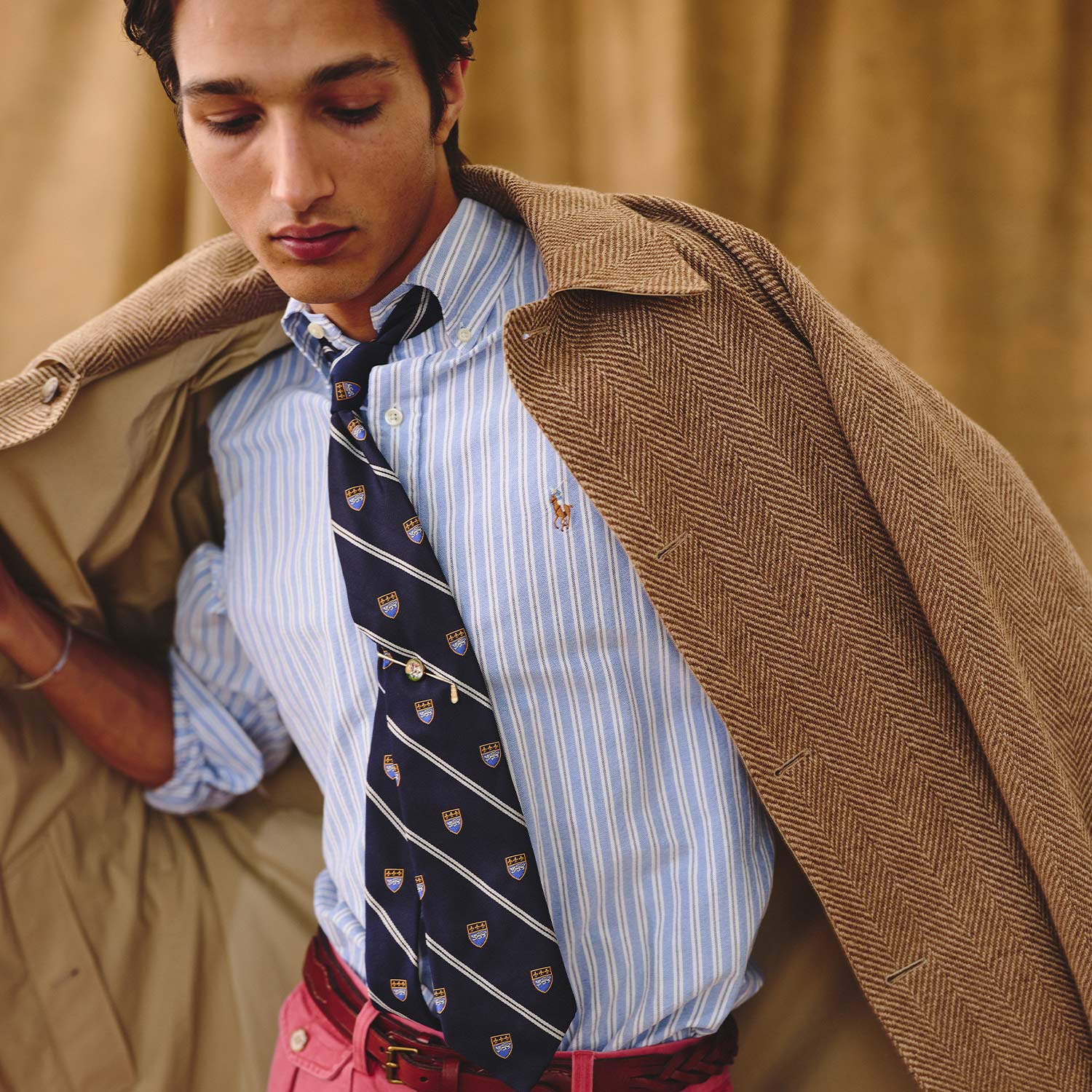 A person wearing a brown coat draped over shoulders, a striped shirt with a tie, and red pants, posing against a neutral background.