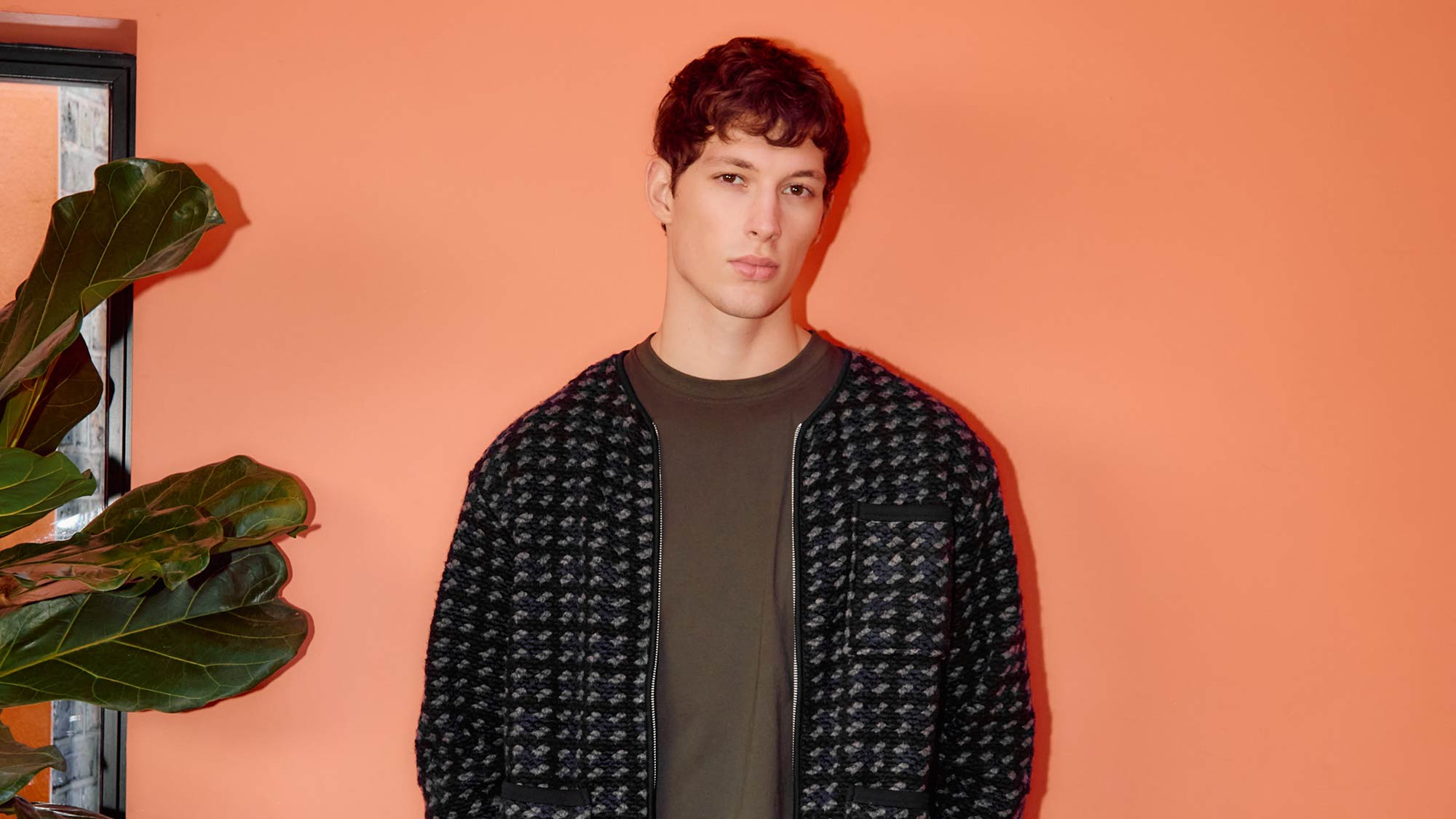 Person in a patterned jacket stands against an orange wall next to a plant.