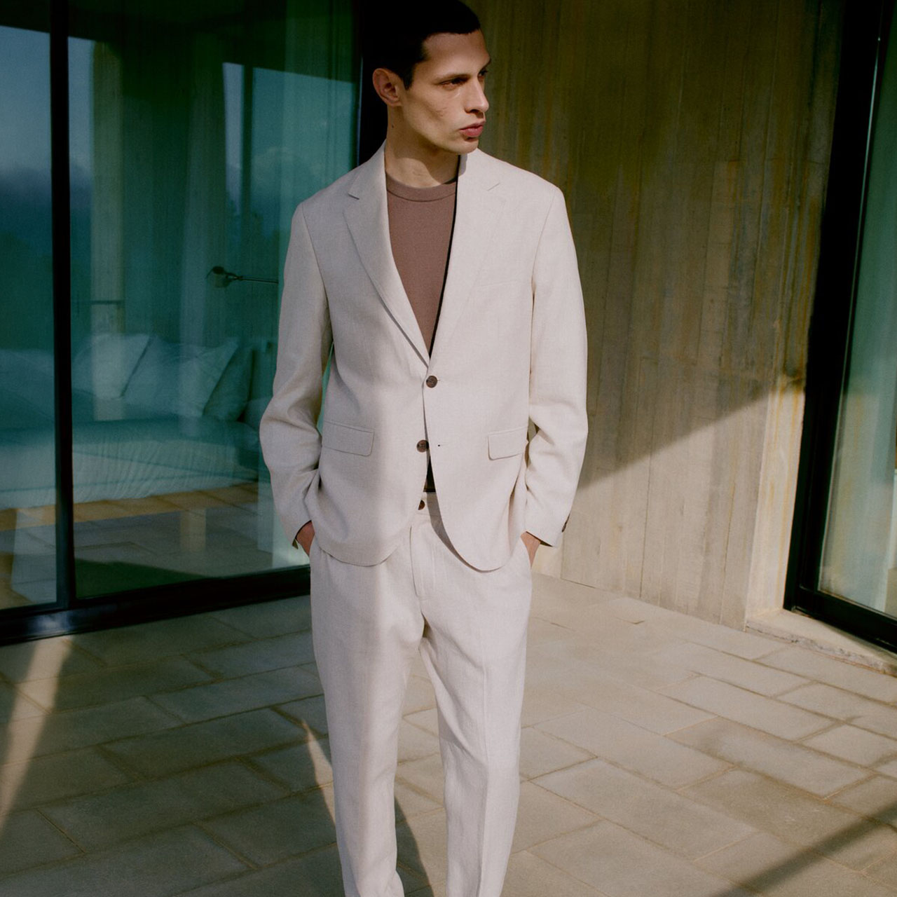 Man wearing cream tailored suit with brown t-shirt, standing in modern interior setting