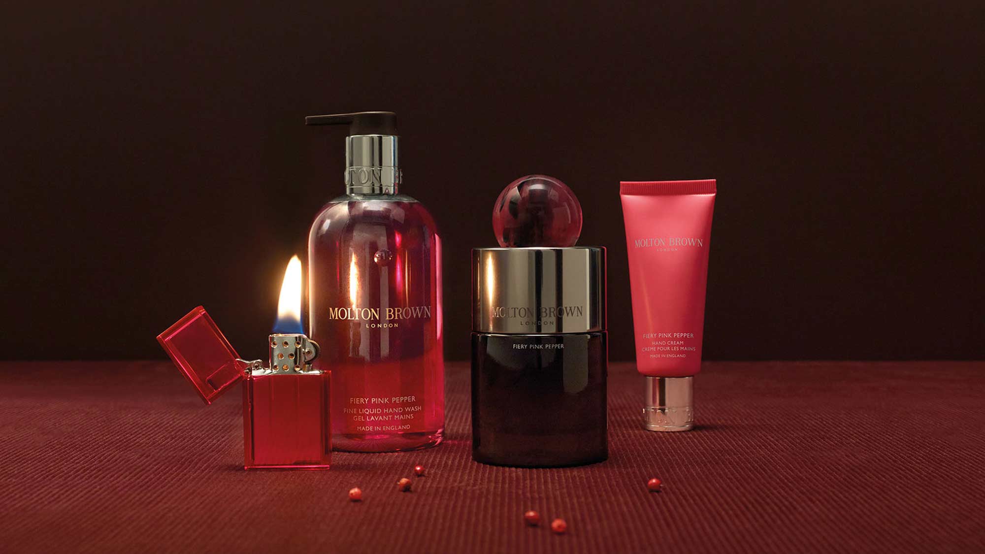 A collection of red-themed cosmetic bottles and a lighter with a flame on a dark red surface.