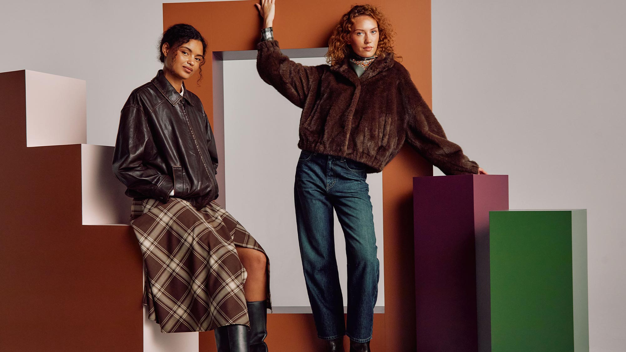 Two women modelling new fashion pieces including brown leather jacket, checked skirt, brown fur jacket and denim jeans against geometric coloured background