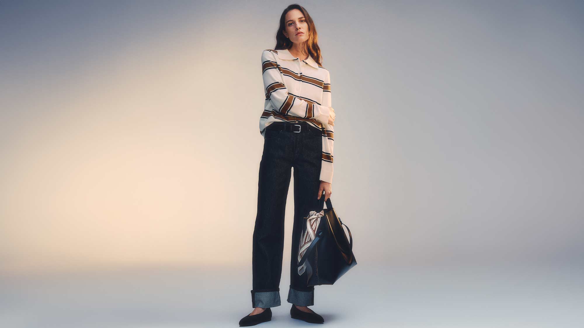 Woman in a striped shirt and dark pants holding a bag against a plain background.