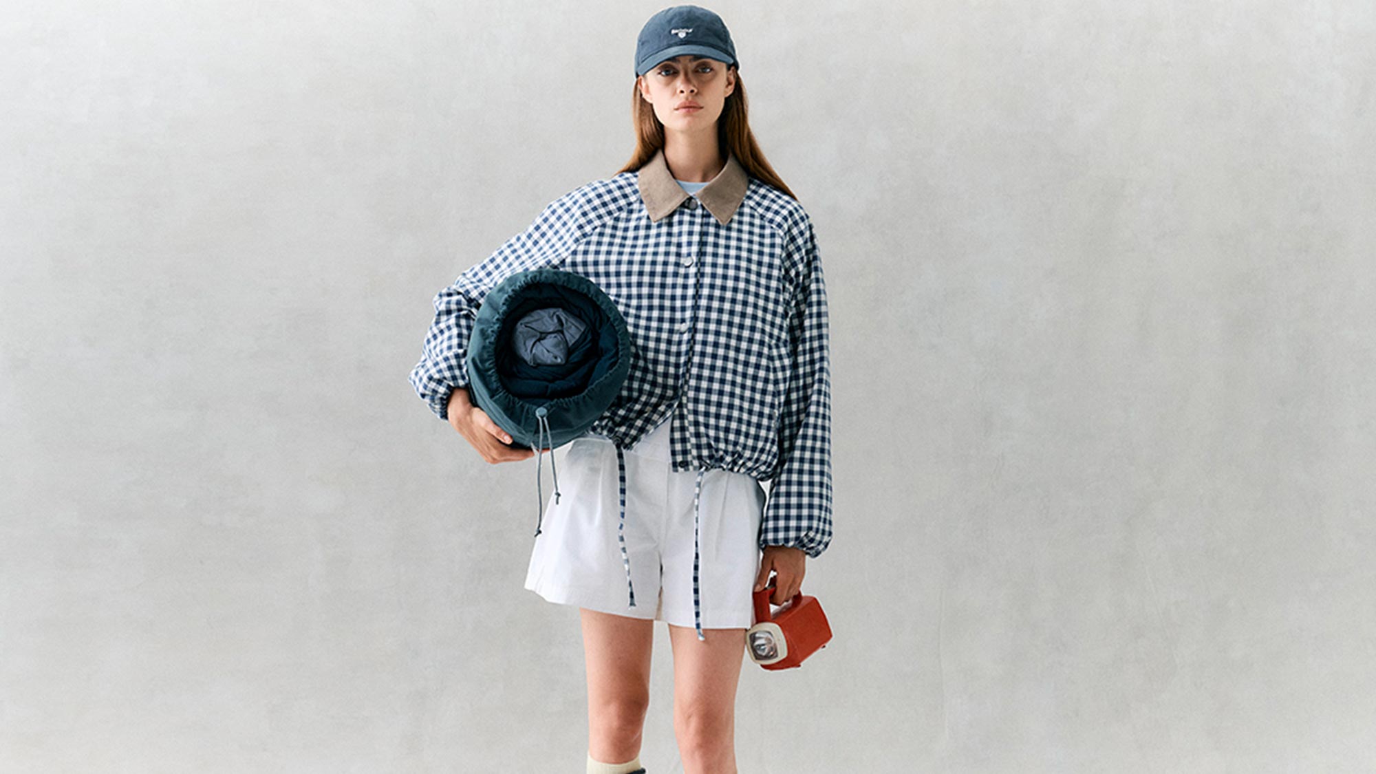 Woman wearing blue and white gingham shirt with white skirt, blue bucket hat, holding blue puffer jacket and red leather bag