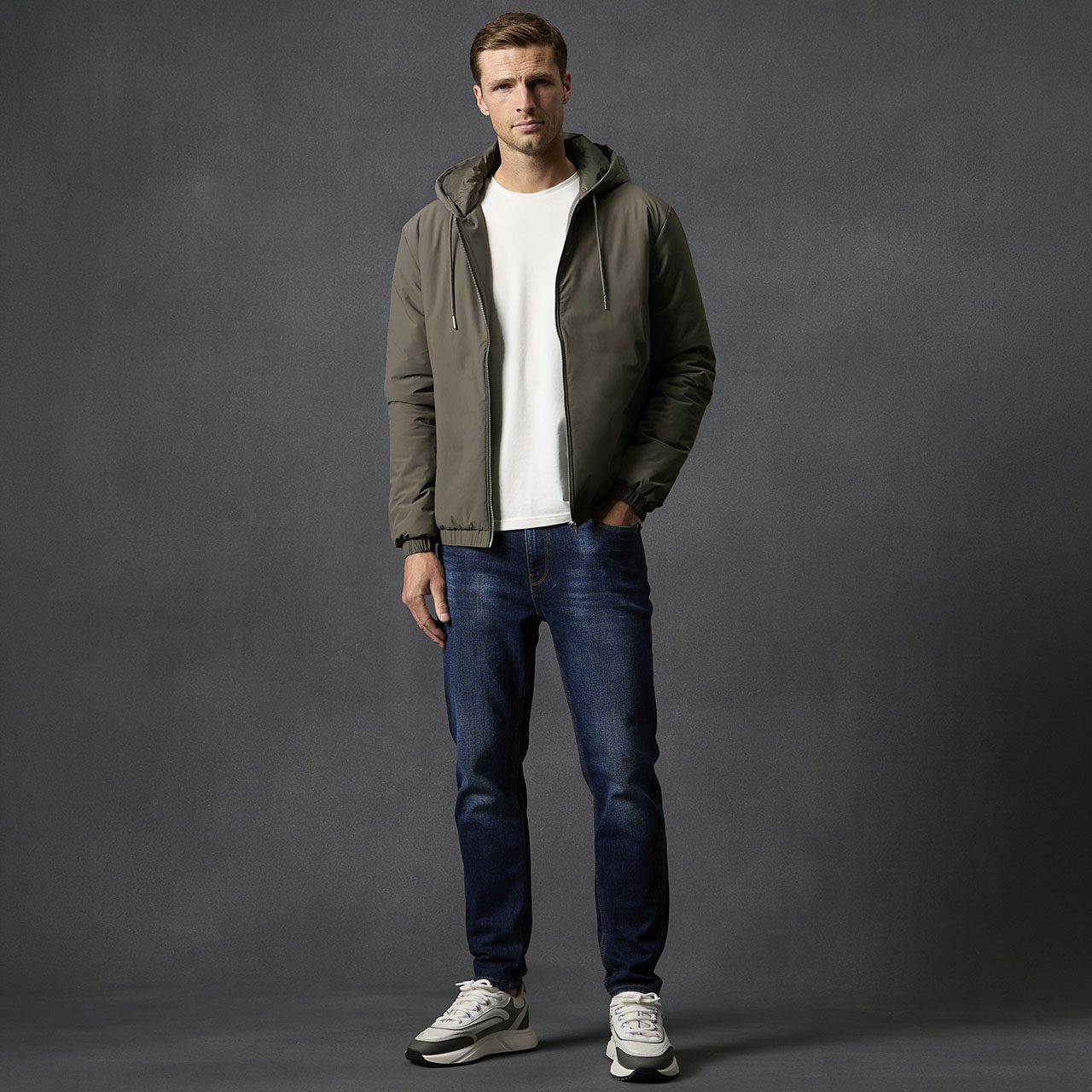 Man wearing olive-green hooded jacket over white t-shirt and blue denim jeans against grey background