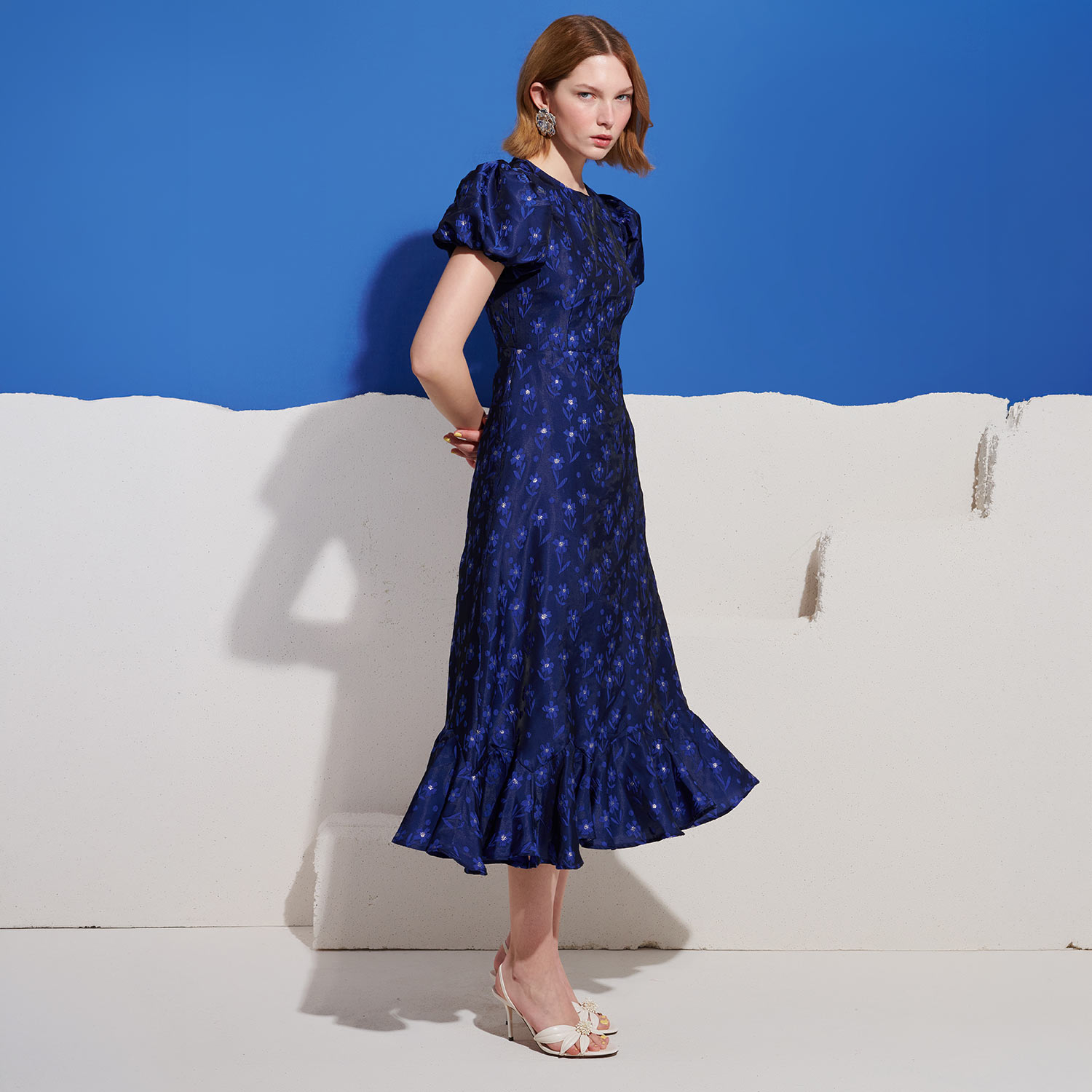 Woman wearing navy blue midi dress with small floral pattern and white strappy heels, standing against white and blue backdrop