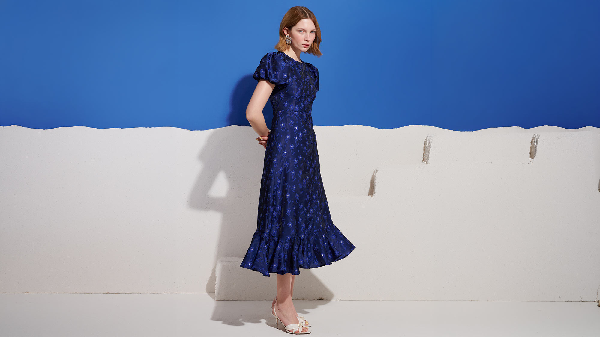 Woman wearing navy blue midi dress with small floral pattern and white strappy heels, standing against white and blue backdrop