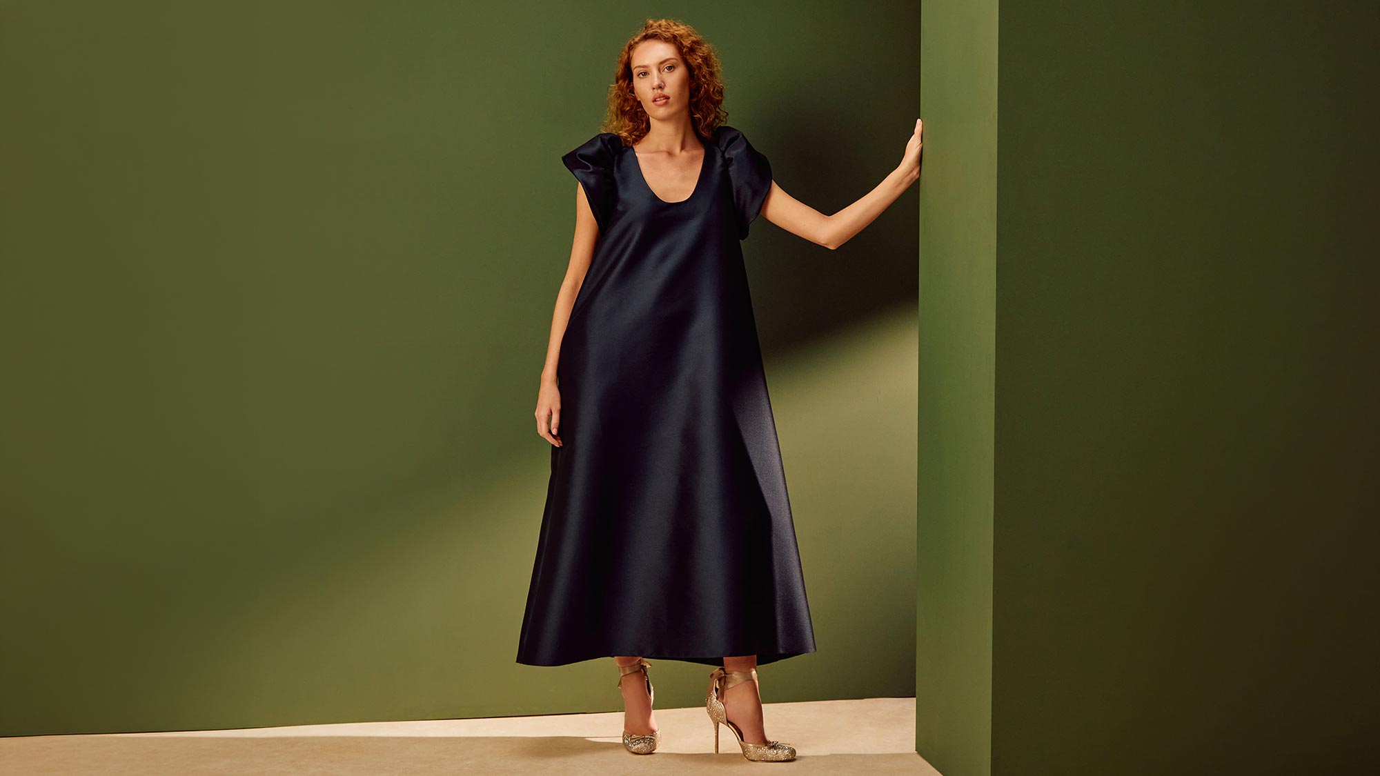 Woman wearing navy A-line midi dress with cap sleeves and metallic heels against green and beige backdrop