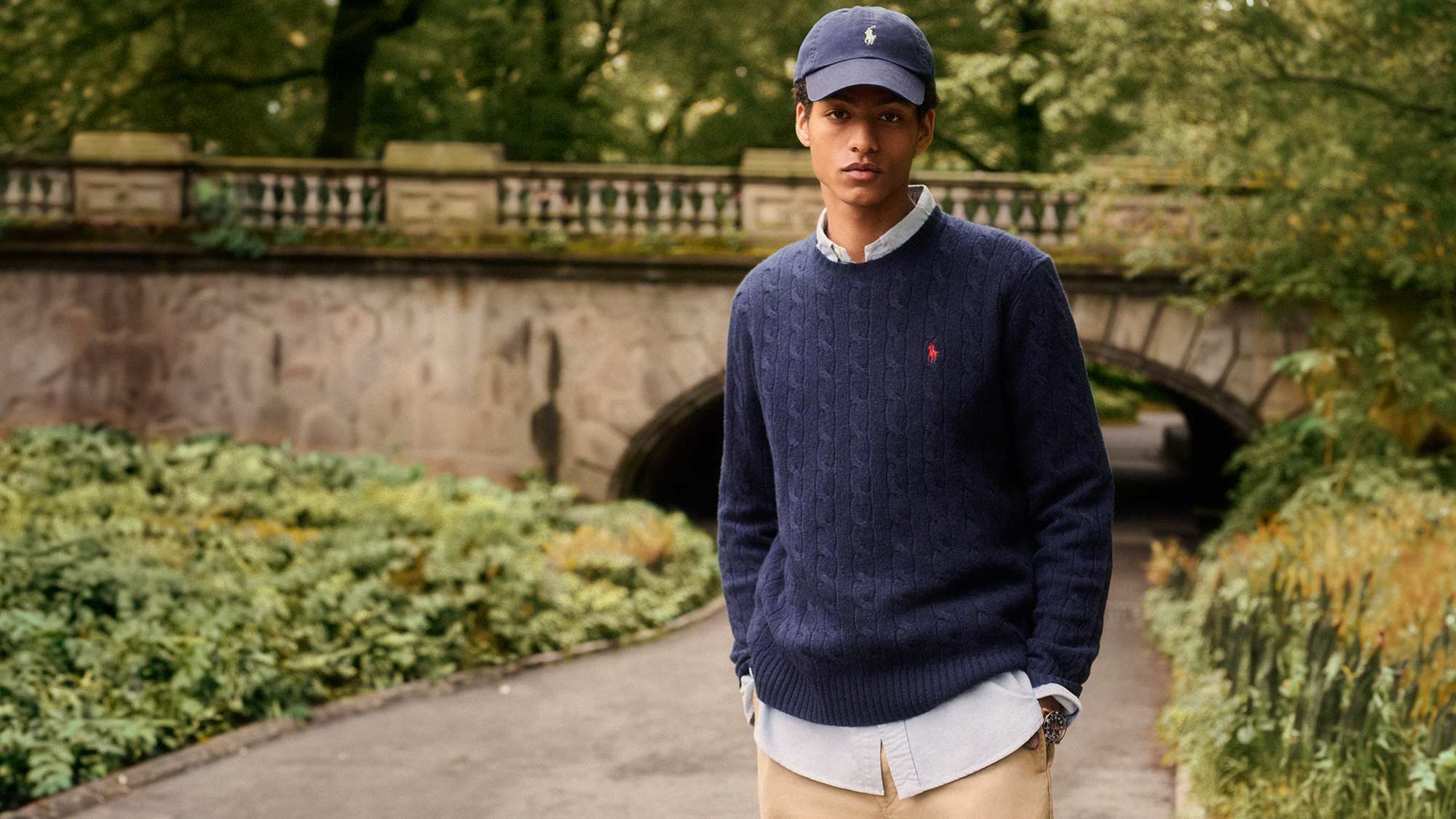 A person in casual attire stands on a path with a stone bridge and lush greenery in the background.