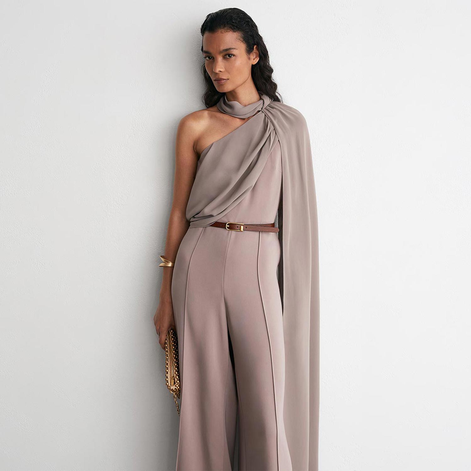 Woman wearing taupe draped one-shoulder dress with brown belt and gold chain bag