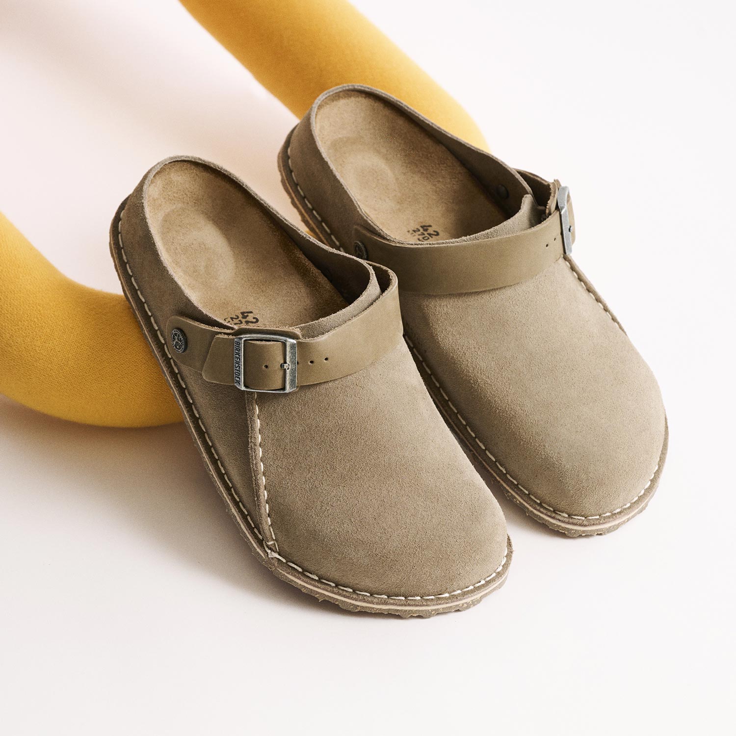 Pair of beige suede clogs with buckle straps and stitched detailing on a yellow curved background