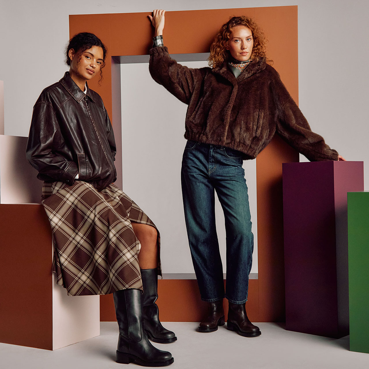 Two women showcasing autumn fashion: one in black leather jacket and tartan skirt, other in brown fuzzy jacket and blue jeans with black boots, positioned among coloured geometric shapes